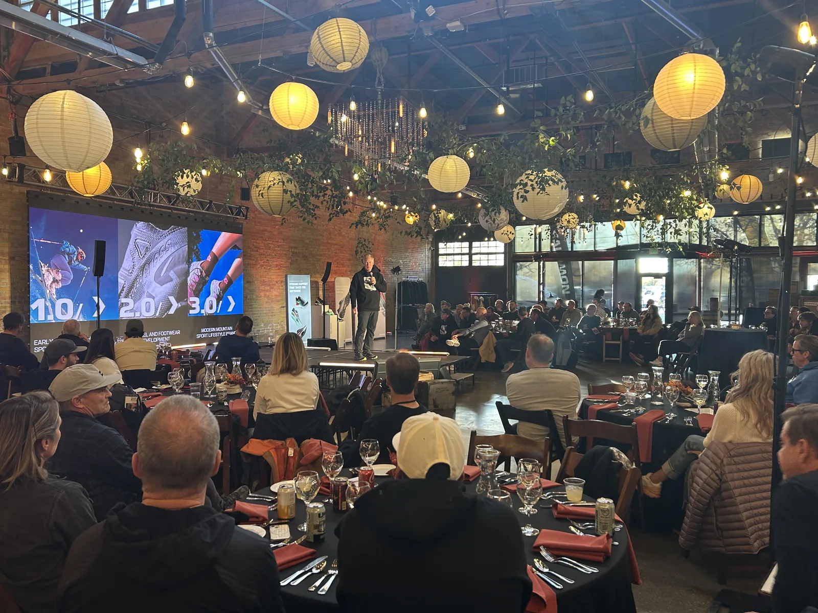 Corporate AV in a brick warehouse venue with a large presentation screen, truss, and hanging lanterns over dinner tables